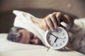 Sleepy young man covering ears with pillow as he looks at alarm clock