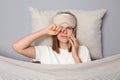 Sleepy young woman wearing white T-shirt and sleep eye mask lies under blanket in bedroom isolated on gray background talking on Royalty Free Stock Photo