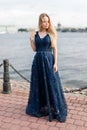 Slender blonde girl in elegant dark-blue floor-length dress on embankment mysteriously looked down Royalty Free Stock Photo