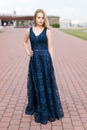 Slender blonde girl in elegant dark-blue floor-length dress on the pavement Royalty Free Stock Photo