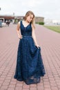 Slender blonde girl touching hem of elegant dark-blue floor-length dress on the pavement Royalty Free Stock Photo