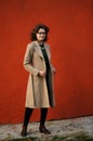 Slender brunette girl in beige coat with spectacles at maroon wa