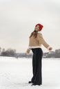 Slender girl in stylish black jeans and beige sheepskin coat with red knitted hat walks in snow outdoors
