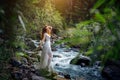 Slender long-haired brunette in white dress posing against small mountain river and green trees. Beautiful young woman walking Royalty Free Stock Photo