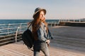 Slender long-haired girl in a denim suit strolls along the sea wharf, looking over her shoulder. Adorable brunette young Royalty Free Stock Photo