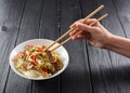 Slender woman hand with chopsticks reaching for Asian udon noodles with meat and vegetables on black background copyspace. Minima