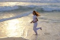 Slender woman in a white tracksuit does a morning run along the edge of the waves on a sandy beach against the background of sunri
