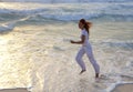 Slender woman in a white tracksuit does a morning run along the edge of the waves on a sandy beach against the background of sunri