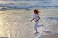 Slender woman in a white tracksuit does a morning run along the edge of the waves on a sandy beach against the background of sunri