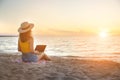 Slender young woman on the beach at the sunset time by the sea works on her laptop holding him on her lap. Freelance Royalty Free Stock Photo