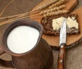 A slice of buttered bread on a wooden board, a crock of milk and mature ears