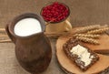 A slice of buttered bread on a wooden board, a crock of milk, a metal cup of red currants and mature ears