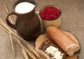 A slice of buttered bread on a wooden board, a crock of milk, a metal cup of red currants and mature ears