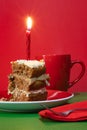 Slice of carrot cake on plate with lighted red candle on red background, red cup and fork. Vertical
