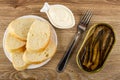 Slice of bread in plate, bowl with mayonnaise, fork, opened jar with sprats on wooden table. Top view