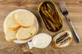 Slice of bread in plate, jar with sprats, bowl with mayonnaise, sandwich with sprats, fork on wooden table. Top view