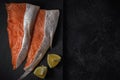Sliced fresh raw salmon lying sideways on a stone cutting board with lemon wedges on a black textured concrete background. top
