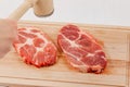 Sliced raw pork steaks meat on a cutting kitchen board on a white wooden table. above them a hammer for beating meat Royalty Free Stock Photo