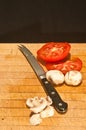 Sliced tomatoes and mushrooms, and whole mushrooms with a serrated bladed knife on wood cutting board