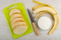 Slices of bread on cutting board, knife, peeled banana, bowl with soft cottage cheese on wooden table. Top view