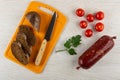 Slices of bread, knife on cutting board, tomato, parsley, smoked sausage in polyethylene pack on table. Top view Royalty Free Stock Photo