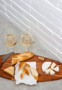 Slices of camembert cheese, baguette and two glasses of white wine on wooden board. White brick wall as background Royalty Free Stock Photo