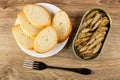 Slices of bread, fork, opened jar with canned sprats on wooden table. Top view