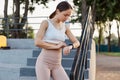 Slim attractive female wearing white top and beige leggins, standing on stairs and looking at her fitness bands, counts steps for