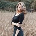 Slim beautiful european young woman in fashionable black t-shirt in vintage blue jeans poses outdoors in a field among dry grass.