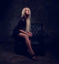 slim blod model with long legs in high heels sitting in fashion armchair in black dress and posing on dark dramatic