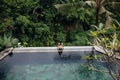 Slim brunette woman in swimsuit relaxing on edge infinity pool in jungle. Palms around and crystal clean water Royalty Free Stock Photo