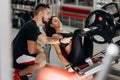 Slim dark-haired girl dressed in black sport clothes is lifting weights with her legs in the gym under the supervision