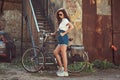 Slim girl in short denim shorts, white t-shirt and sunglasses, posing with the city bicycle near an old abandoned brick Royalty Free Stock Photo