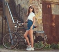 Slim girl in short denim shorts, white t-shirt and sunglasses, posing with the city bicycle near an old abandoned brick Royalty Free Stock Photo