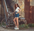 Slim girl in short denim shorts, white t-shirt and sunglasses, posing with the city bicycle near an old abandoned brick Royalty Free Stock Photo