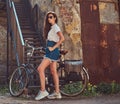 Slim girl in short denim shorts, white t-shirt and sunglasses, posing with the city bicycle near an old abandoned brick Royalty Free Stock Photo