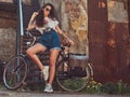 Slim girl in short denim shorts, white t-shirt and sunglasses, posing with the city bicycle near an old abandoned brick Royalty Free Stock Photo