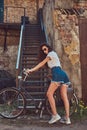 Slim girl in short denim shorts, white t-shirt and sunglasses, posing with the city bicycle near an old abandoned brick Royalty Free Stock Photo