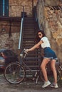 Slim girl in short denim shorts, white t-shirt and sunglasses, posing with the city bicycle near an old abandoned brick Royalty Free Stock Photo