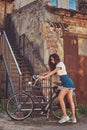 Slim girl in short denim shorts, white t-shirt and sunglasses, posing with the city bicycle near an old abandoned brick Royalty Free Stock Photo