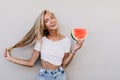 Slim glad girl touching her blonde hair and smiling. Studio portrait of pretty lady with watermelon