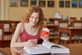 Slim good looking young student sitting in local library, holding papercup of coffee and smartphone, looking at mobile screen,