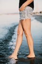 Slim legs of a young and beautiful girl in shorts sit on the sand in the waves
