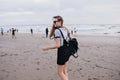 Slim pale girl with black backpack walking around beach with happy smile. Outdoor shot of magnificent lady having fun