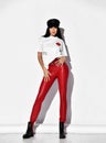 Young sensual woman vamp in tight red leather pants, white t-shirt and leather cap stands holding hands on her waist Royalty Free Stock Photo