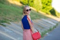 Slim tall blonde girl in blue top and light pink skirt wearing sunglasses with a small red shoulder bag to look back. rear view.