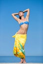 Slim tall girl with yellow sarong on hips standing with arms raised against sky