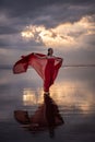 Slim woman dancing on the beach during sunset. Water reflection. Pretty Caucasian woman wearing long red dress. Perfect body shape
