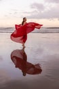 Slim woman dancing on the beach during sunset. Water reflection. Pretty Caucasian woman wearing long red dress. Perfect body shape