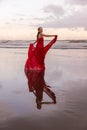 Slim woman dancing on the beach during sunset. Water reflection. Pretty Caucasian woman wearing long red dress. Perfect body shape